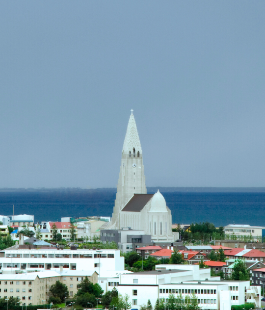A panoramic view of downtown Reykjavík featuring Hallgrímskirkja, a tall white church with a pointed tower. The city is surrounded by colorful houses and green trees. In the background, the b