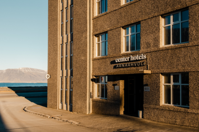 Entrance to Center Hotels Arnarhvoll in Reykjavík, with warm sunlight on the façade and the sea and mountains in the background.