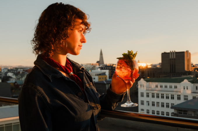 Guest holding a cocktail at SKÝ Lounge & Bar in golden sunlight, with Reykjavík’s rooftops and Hallgrímskirkja in the background.