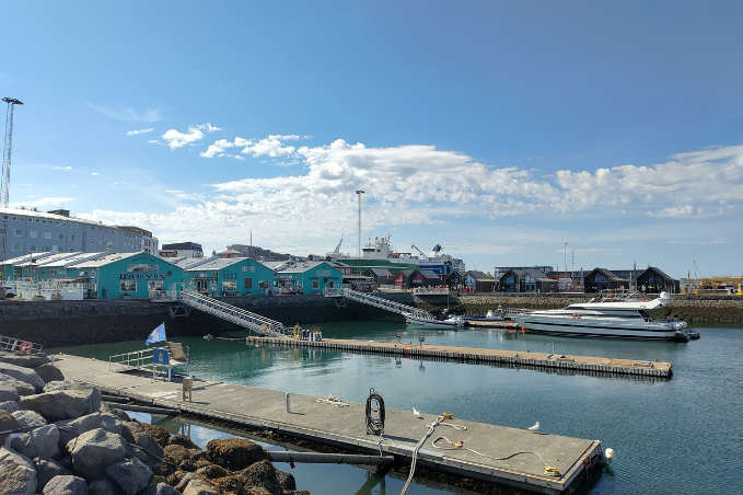 Boats and floating docks at Grandi harbour in Reykjavík, with turquoise waterfront buildings under a blue sky.