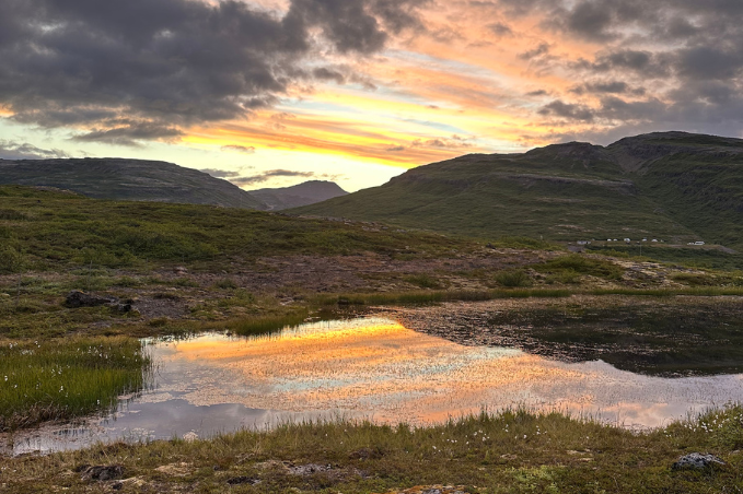 Golden midnight sun colours reflected in a small pond, surrounded by green hills beneath dramatic clouds in Iceland.