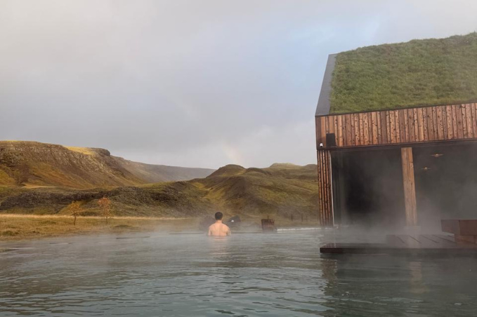 A guest enjoys a steaming outdoor pool overlooking grassy Icelandic hills beside a timber building with a turf roof.