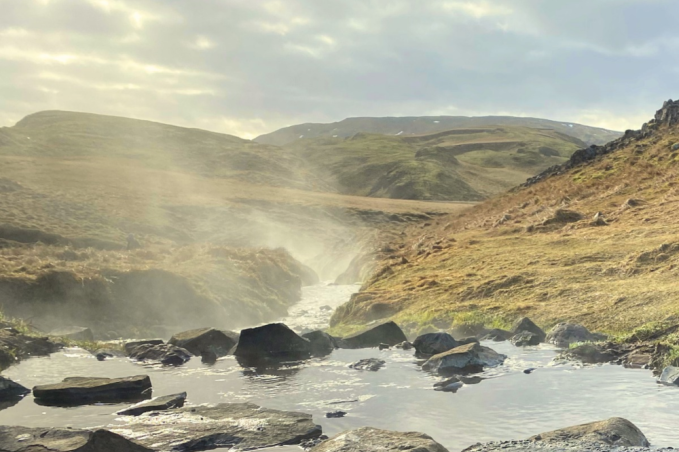 Steam rises from a rocky stream winding between golden grassy slopes and rugged hills in Iceland.