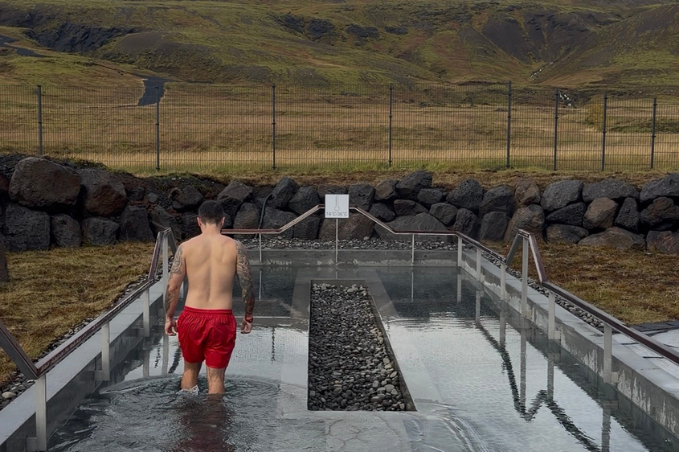 A guest in red swimming shorts steps into an outdoor plunge pool bordered by dark volcanic rocks, with grassy hills beyond.