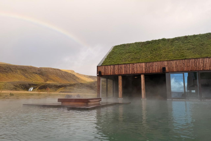 Steam rises from an outdoor pool beside a timber spa building with a turf roof, with a rainbow over the surrounding hills.