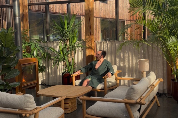 A guest in a green bathrobe relaxes beside a fireplace in a sunlit spa lounge with tropical plants and wooden furniture.
