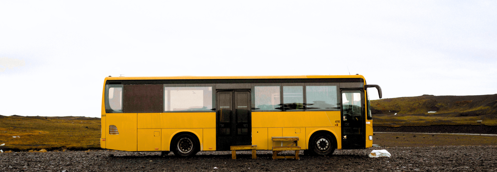 A yellow bus is parked alone on a gravel area in the Icelandic highlands. A simple wooden bench sits in front of it, and the surrounding landscape is vast and barren, with low mossy hills und