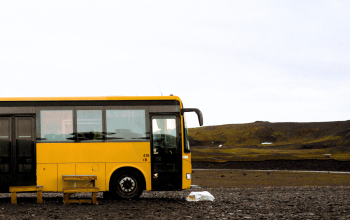 A yellow bus is parked alone on a gravel area in the Icelandic highlands. A simple wooden bench sits in front of it, and the surrounding landscape is vast and barren, with low mossy hills und