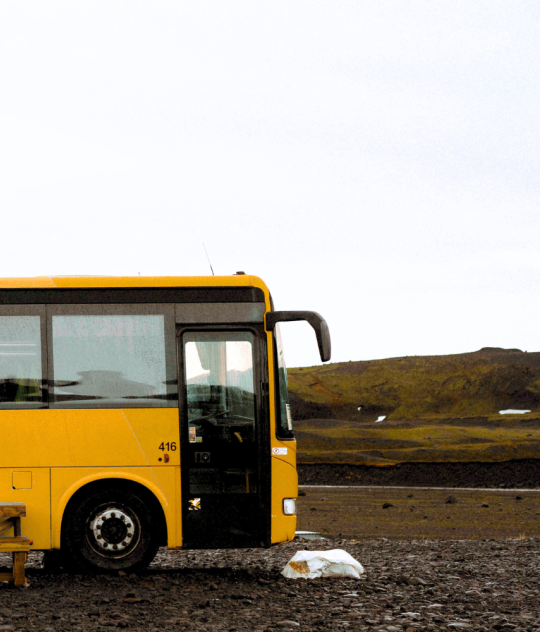 A yellow bus is parked alone on a gravel area in the Icelandic highlands. A simple wooden bench sits in front of it, and the surrounding landscape is vast and barren, with low mossy hills und