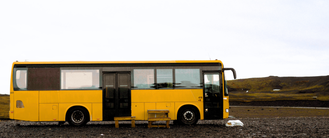 A yellow bus is parked alone on a gravel area in the Icelandic highlands. A simple wooden bench sits in front of it, and the surrounding landscape is vast and barren, with low mossy hills und