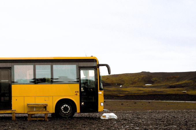 A yellow bus is parked alone on a gravel area in the Icelandic highlands. A simple wooden bench sits in front of it, and the surrounding landscape is vast and barren, with low mossy hills und