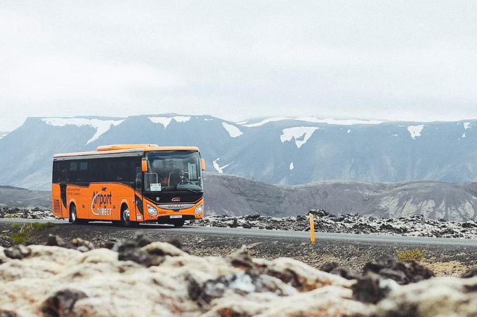 Orange airport shuttle bus driving along a road with Icelandic mountains in the bakcground.