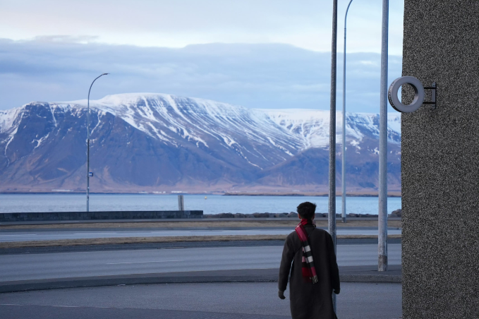 Person standing outside Center Hotels Arnarhvoll near the harbor looking toward snow-covered mountains across the water.