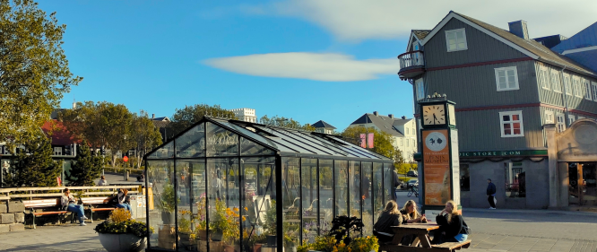 Sunny day in downtown Reykjavik with a small glass greenhouse, benches, and people relaxing near a clock tower.