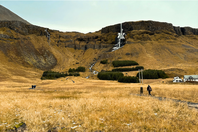 Two people walking through a wide valley toward a waterfall in rural Iceland