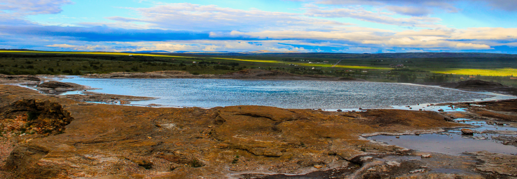 A calm geothermal hot spring surrounded by rocky terrain with green fields and a cloudy sky in the distance.