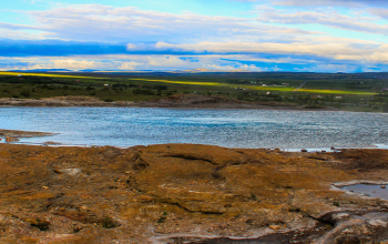 A calm geothermal hot spring surrounded by rocky terrain with green fields and a cloudy sky in the distance.