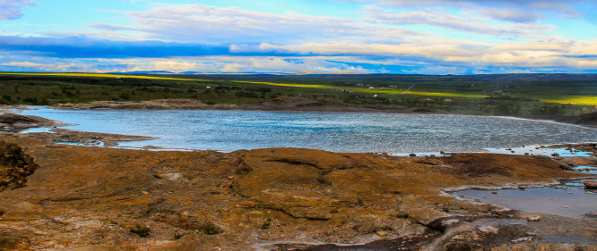 A calm geothermal hot spring surrounded by rocky terrain with green fields and a cloudy sky in the distance.