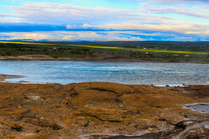 A calm geothermal hot spring surrounded by rocky terrain with green fields and a cloudy sky in the distance.