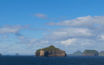 A wide-angle view of the Westman Islands in Iceland, with dark blue ocean in the foreground and scattered green islands under a bright blue summer sky with light clouds. A few seabirds are fl