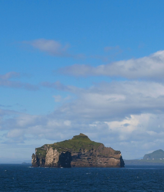 A wide-angle view of the Westman Islands in Iceland, with dark blue ocean in the foreground and scattered green islands under a bright blue summer sky with light clouds. A few seabirds are fl