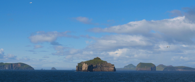 A wide-angle view of the Westman Islands in Iceland, with dark blue ocean in the foreground and scattered green islands under a bright blue summer sky with light clouds. A few seabirds are fl