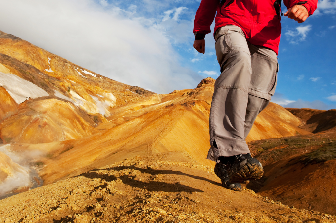 Close-up of a hiker wearing sturdy boots, beige trousers and a red jacket on an ochre mountain trail.