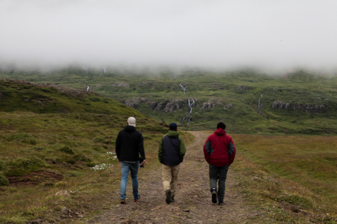 Three hikers in outdoor jackets follow a gravel trail towards misty green hills and small waterfalls.