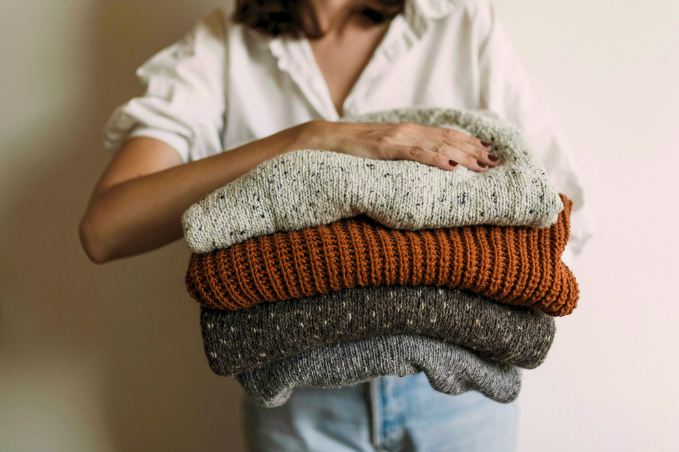 Person holding a stack of folded knitted sweaters in cream, rust, charcoal and grey.