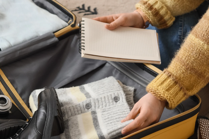 Person packing a yellow suitcase with black ankle boots, a knitted cardigan and a camera while holding a notebook.