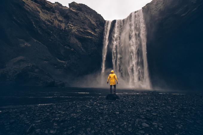 Person in a yellow waterproof jacket facing a large waterfall surrounded by dark cliffs and rocky ground.
