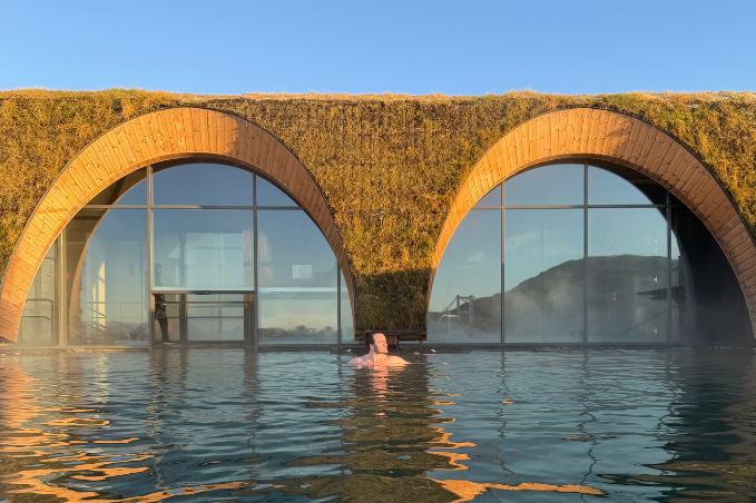 Person bathing in an outdoor geothermal pool at Laugarás Lagoon with modern arched buildings and steam rising in Iceland