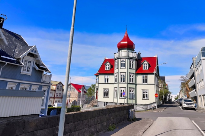 Historic Reykjavík house with pale green trim, a bright red roof and a turret, beside a blue house under a clear sky.